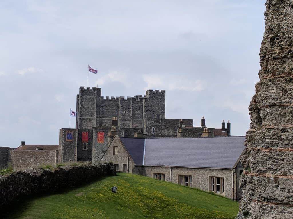 In front is the Church of St. Mary-in-Castro next to the Roman lighthouse. In the background is Dover Castle, one of the largest castles in England. A British flag flies above the castle.