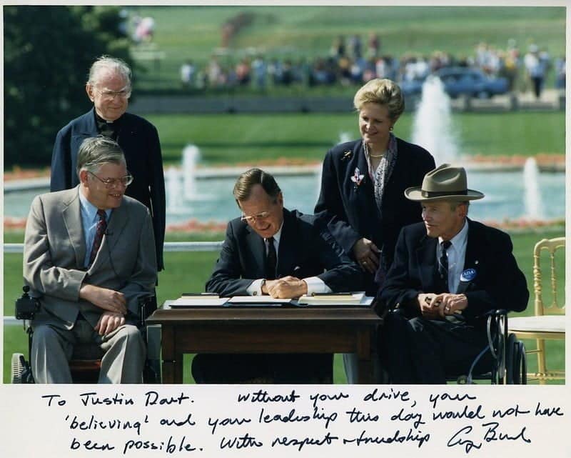 George Bush, seated a table in front of a fountain and pond, perhaps in D.C., signs the Americans with Disabilities Act between four other people.